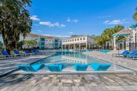 Luxurious apartment complex featuring a stunning swimming pool with modern lounge chairs, surrounded by palm trees and a clear blue sky.