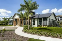 Luxurious modern clubhouse surrounded by lush landscaping and palm trees, featuring a sleek metal roof and stone columns.