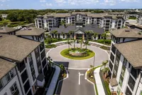 Aerial view of a modern apartment complex with lush landscaping, palm trees, and a stylish central roundabout, showcasing elegant architecture and spacious living options.