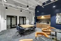 Modern shared office space with sleek wooden table, stylish blue chairs, elegant kitchenette, and chic lounge area.