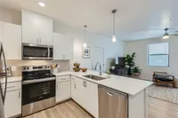 Three-BR Apartments in West Phoenix, AZ - Meritum Sheely Farms - Kitchen with Vinyl Plank Flooring, Quartz Counters, Stainless Steel Appliances, White Cabinets, a Kitchen Island, Modern Light Fixtures, a Ceiling Fan, the Living Room, and Stylish Decor