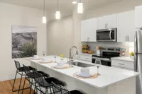 Three-BR Apartments in West Phoenix, AZ - Meritum Sheely Farms - Kitchen with Vinyl Plank Flooring, Quartz Counters, Stainless Steel Appliances, White Cabinets, a Kitchen Island, Modern Light Fixtures, a Ceiling Fan, the Living Room, and Stylish Decor