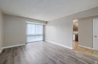 Bright and inviting living room with elegant wood flooring, large window with vertical blinds, and a glimpse of a modern kitchen.