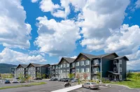 Modern apartment complex with sleek design set against a backdrop of picturesque clouds, offering ample parking and landscaped surroundings.