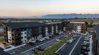 Modern apartment complex surrounded by scenic mountains at sunset, featuring ample parking and stylish architecture.