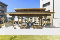 Modern outdoor patio area with wooden pergola, picnic tables, and built-in grills, surrounded by stylish apartment buildings.