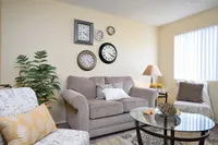 Camelot Apartment's inviting living room with a cozy beige sofa, elegant wall clock display, and abundant natural light streaming through vertical blinds.