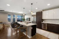 Sugarmont Townhome Kitchen View of the two bedroom townhouse at Sugarmont Apartments, pendant lighting, showing ceiling fan, island kitchen and quartz countertops with two tone cabinets and upgraded appliances, grey suede bar stools, dining table with white chairs, wine fridge, and grey couch with large windows showing mountain views.