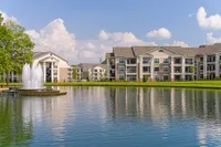 One, Two, and Three Bedroom Apartments in Durham, NC Luxurious apartment complex, Heights at Meridian, overlooking a serene pond with a fountain, surrounded by lush greenery under a bright blue sky.