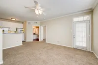 Bright and airy living room with plush carpet, ceiling fan, and an open kitchen with white cabinetry at Stonebriar of Frisco Apartments.