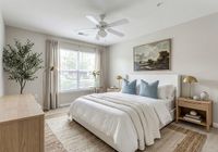 Bright and serene bedroom featuring a cozy bed with plush pillows, elegant decor, and natural light streaming through a large window at Stratford at Hillcrest Apartments