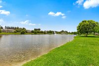 Serene lake view with lush green grass, vibrant trees, and elegant homes under a bright blue sky.