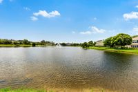 Serene lakeside view with a charming fountain, surrounded by lush greenery and elegant homes under a bright blue sky.