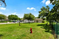 Vibrant dog park with lush green grass, agility equipment, and a bright red fire hydrant, surrounded by trees and secure fencing, under a clear blue sky.