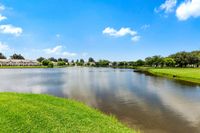 Serene lakeside view with charming townhouses, lush greenery, and a sparkling fountain under a clear blue sky.