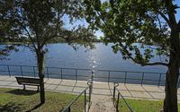 Lakeside walkway with a bench, iron railing, and stairway under tree shade overlooking calm water.