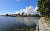Curving lakeside path with black railing, tree shade, and expansive water views under a blue sky.
