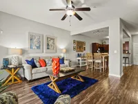 Bright apartment living room with plush sofa, colorful cushions, wood flooring, and open kitchen layout.