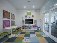 Vibrant playroom with colorful geometric carpet, modern furniture, artistic wall decor, and glass doors leading to a bright fitness area.