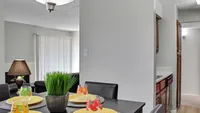 Bright dining area with modern table setting, lush plant centerpiece, and cozy seating, leading into a functional kitchen space.
