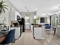 Bright, open-concept living space featuring a modern kitchen with sleek black appliances, a cozy seating area, and elegant dining setup, enhanced by large windows and stylish decor.