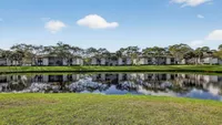 Serene waterfront residences reflecting in a tranquil pond, surrounded by lush greenery and clear blue skies.