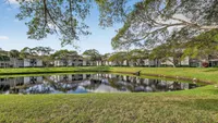 Serene apartment complex with lush greenery and a tranquil pond reflecting elegant residences amidst tall trees under a clear blue sky.