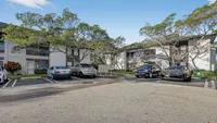 Charming apartment complex with lush trees and ample parking, offering serene living under a bright blue sky.