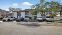 Charming suburban apartment building with a lush landscape, ample parking, and inviting balconies on a sunny day.