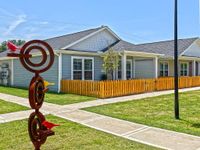 Charming single-story home with pristine gray siding, cheerful yellow fence, and bold artistic sculpture in a lush green setting.