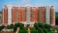 Impressive high-rise apartment building with elegant red and white facade, featuring numerous balconies and surrounded by lush green landscaping.