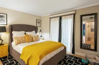 Cozy bedroom with elegant decor, featuring a luxurious bed with vibrant yellow accents, modern art, and a large window with soft drapery, creating a serene and inviting atmosphere.