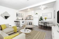 A modern open-plan living space with a beige sofa, round coffee table, dining area, and kitchen. Decor includes yellow accents, a wall mirror, and a ceiling fan. Black bar stools at the counter.