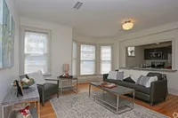 Charming and cozy living room with elegant hardwood flooring, featuring a plush gray sofa, stylish coffee table, and large windows providing ample natural light; perfect open-concept layout flowing into a modern kitchen with stainless steel appliances.