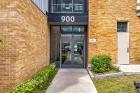 Modern apartment entrance with brick facade and glass double doors, labeled '900', surrounded by well-kept greenery.