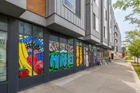 Vibrant urban setting with artistic murals on storefront windows, featuring fresh fruits and bold typography, promising an upcoming market in a contemporary apartment building.