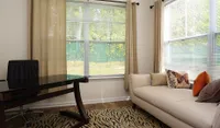Bright and inviting living area with a modern glass desk, cozy beige sofa, and chic animal print rug next to large sunlit windows.