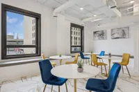 Contemporary dining area with bold blue and yellow chairs, chic round tables, and large windows offering a vibrant city view.
