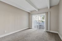 Bright and inviting room with neutral walls and plush carpeting, featuring floor-to-ceiling windows with vertical blinds revealing a garden view.