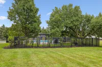 Serene outdoor space with lush green trees, fenced area, and a glimpse of a refreshing swimming pool under a clear blue sky.