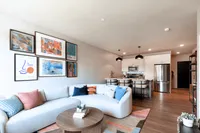 Modern open-concept living room with stylish decor, featuring a chic sectional sofa, vibrant artwork, and a sleek kitchen area with stainless steel appliances.