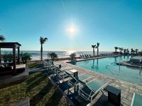 Luxurious beachfront pool area with sun loungers, palm trees, and stunning ocean views under a bright blue sky.