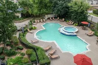 Inviting aerial view of a luxury apartment complex pool area, featuring a uniquely shaped pool with adjacent hot tub, surrounded by lush greenery and stylish lounge chairs.