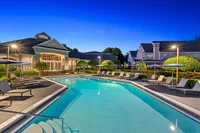 Elegant apartment complex with sparkling swimming pool, surrounded by lush greenery and stylish lounge chairs under a serene evening sky.