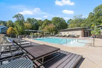 Luxurious outdoor swimming pool area with modern lounge chairs, surrounded by lush green trees and clear blue skies.