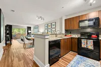 Modern open-concept kitchen and living area with sleek wooden cabinetry, granite countertops, and vibrant decor, leading to a bright seating area with large windows overlooking greenery.
