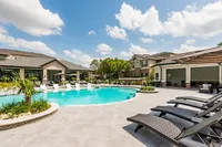 Luxurious apartment complex featuring a sparkling outdoor pool surrounded by elegant lounge seating, lush landscaping, and modern architecture under a bright blue sky.