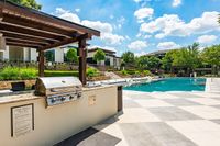 Luxurious outdoor pool area with modern BBQ grill, surrounded by lush greenery and stylish pergolas under a bright blue sky.