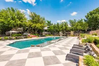 Luxurious apartment complex pool area featuring a sparkling swimming pool, elegant sun loungers, lush greenery, and a stylish checkered patio under a clear blue sky.