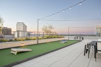 Modern rooftop terrace with comfortable seating, cornhole game, and ambient string lights against a vibrant cityscape.